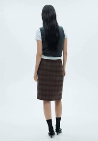 Jupe à carreaux ajustée dans des tons bruns et beiges, mi-longue, associée à un haut blanc à manches courtes et un gilet sombre, portée avec des chaussettes noires et des talons.