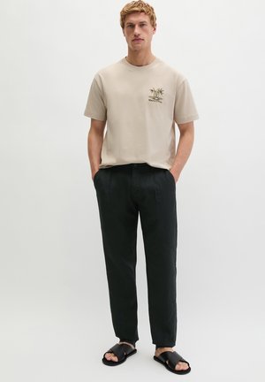 Mann trägt beige kurzärmeliges Hemd mit kleinem Palmenmuster, schwarze Hose und schwarze Sandalen, steht mit den Händen in den Taschen.