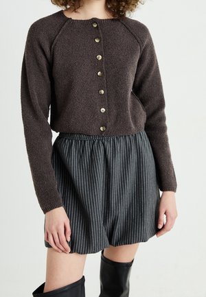 Personne portant un pull boutonné marron foncé, un short à fines rayures noir et blanc, et des bottes noires montantes sur un fond uni.