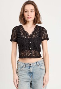 Crop top noir en crochet avec des manches courtes, encolure en V et fermeture à boutons. Présente un motif floral et une texture similaire à de la dentelle. Associé à un jean taille haute.