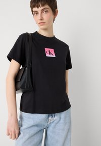 Melnā kokvilnas T-kreklā ar apaļu kakla izgriezumu, īsām piedurknēm un rozā gradienta kvadrātu ar "cK" logotipu. Saskaņots ar gaiši ziliem džinsu biksēm un melnu somu.