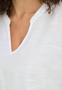 Biała lniana bluzka z luźnym dekoltem w literę V i teksturowaną powierzchnią, koncentrująca się na prostej konstrukcji i lekkim materiale.