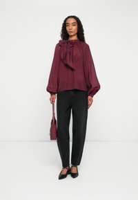 Femme debout portant un chemisier bordeaux avec un nœud, un jean noir, des talons noirs, et tenant un sac à main assorti bordeaux sur fond blanc.