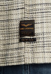 Étiquette en tissu PME Legend noire et dorée cousue sur un vêtement à carreaux beige texturé et noir, au-dessus d’un denim bleu foncé.