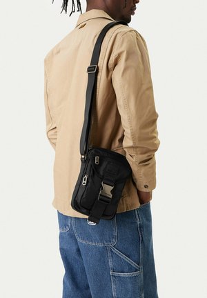 Mężczyzna w beżowej kurtce i niebieskich dżinsach, noszący na ramieniu małą czarną torebkę na pasku z klamrą i detalami na suwaku.