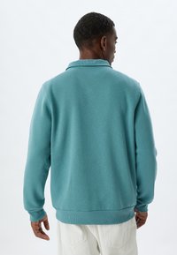 Ljusblå sweatshirt med krage, ribbstickade ärmslut och nederkant. Söt textur, avslappnad design, enfaldig bakvy.