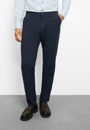 Man draagt marineblauwe broek, lichtblauw overhemd met knopen en bruine leren schoenen, staand met één hand in de zak.