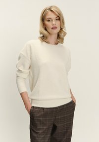 Blondynka z kręconymi włosami, ubrana w biały sweter i brązowe spodnie w kratę, stojąca z rękami w kieszeniach na neutralnym tle.