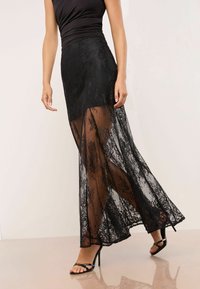 Schwarzer Spitzenrock mit transparenter Überlage, das Blumenmuster zeigt. Enganliegende Taille und ausgestellter Saum, kombiniert mit schwarzen Absatzsandalen.