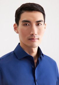 Blauwe overhemd met een spread collar. Gladde textuur, enkele borstknopen, getailleerd ontwerp. Model staat rechtop tegen een neutrale achtergrond.