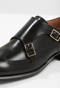 Chaussure habillée en cuir noir avec double bride monk et boucles dorées, coutures détaillées et bout pointu.