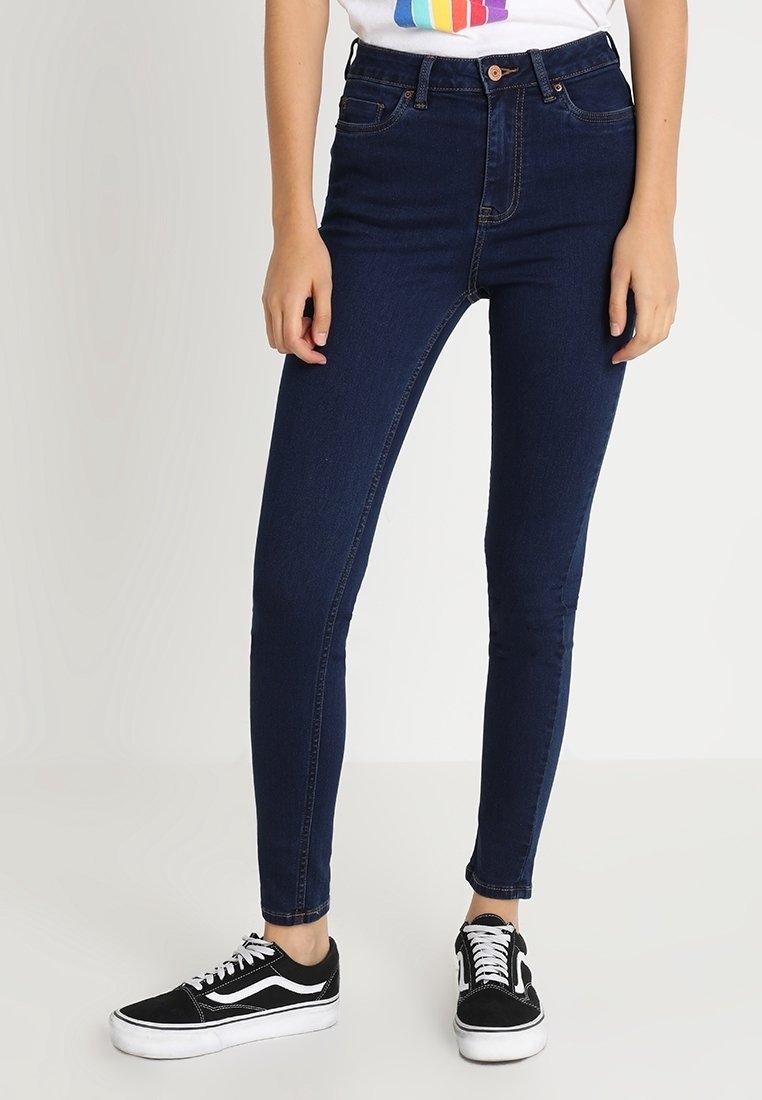 Jeans ajustados de denim azul oscuro, de cintura alta y con una textura suave, combinados con zapatillas negras y blancas, de pie contra un fondo liso.