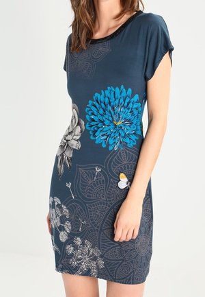 Femme portant une robe bleu foncé à manches courtes avec de grands motifs floraux et papillons bleus, gris et blancs.
