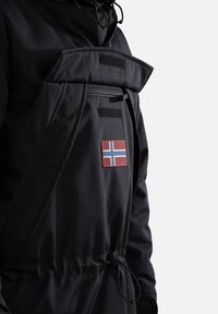 Veste noire avec poche poitrine zippée, rabat à velcro, taille réglable par cordon de serrage, et un patch croix nordique rouge et bleu sur le devant.