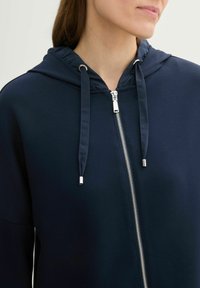 Hoodie zippé bleu marine en tissu doux, avec capuche à cordon, détails de fermeture éclair argentés et design décontracté avec un minimum de coutures.