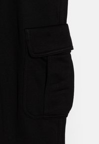 Mustade kaubapükste detail, millel on suur klapiga tasku küljes, valmistatud pehmest, tekstuuriga kangast, tugevdavate õmblustega.