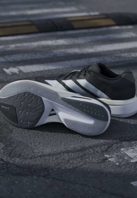 Paire de chaussures de course Adidas noires et blanches, l’une montrant une semelle Lightstrike portant l’inscription Adizero, posée sur l’asphalte près d’un passage piéton.