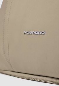 Produit en cuir beige clair avec une texture lisse, présentant un logo argenté embossé et des coutures visibles le long du bord.