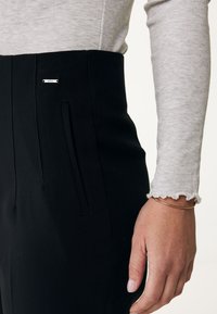 Svarta skräddarsydda byxor i en slät tygkvalitet, med en diskret ficka och en liten metallikon i närheten av midjan.