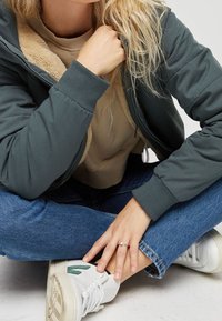 Grüne Kapuzenjacke mit beigem Sherpa-Futter, kombiniert mit einem beigen Pullover. Jeans und weiße Sneakers mit grünen Akzentdetails.