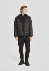 Giacca a vento nera con cappuccio, zip frontale e texture trapuntata, abbinata a pantaloni della tuta neri e sneakers.