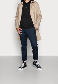 Beige vattentät trenchcoat över en svart hoodie, marinblå byxor och svarta högskaftade sneakers med vita sulor. Avslappnad stadsoutfit.