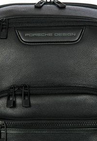 Schwarzer Leder-Rucksack mit strukturiertem Oberflächenmaterial, ausgestattet mit zwei Reißverschlussfächern und einem auffälligen geprägten Etikett mit der Aufschrift "Porsche Design."