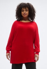 Κόκκινο πουλόβερ με μακριά μανίκια, ριμπ υφή, στρογγυλή λαιμόκοψη και άνετη εφαρμογή, σε συνδυασμό με μαύρο παντελόνι. Λείο ύφασμα και casual σχεδίαση.