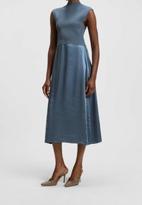 Modrá midi šaty s přiléhavým živůtkem, bez rukávů a volnou sukní z hladkého saténového materiálu. Nosené s béžovými vysokými podpatky.