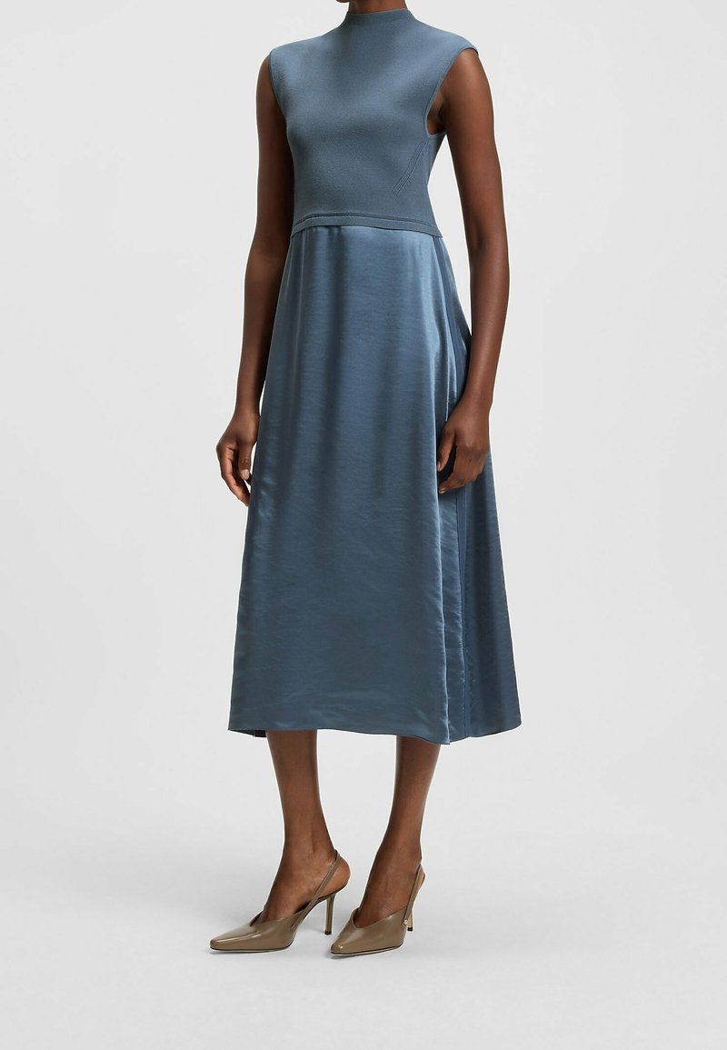 Modrá midi šaty s přiléhavým živůtkem, bez rukávů a volnou sukní z hladkého saténového materiálu. Nosené s béžovými vysokými podpatky.