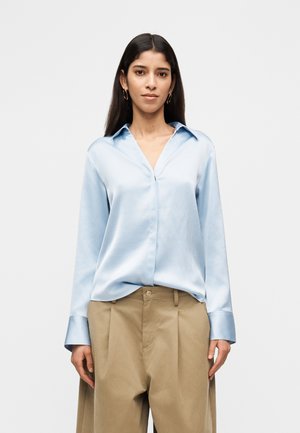 Hosszú sötét hajú nő arany karika fülbevalóval, világoskék szatén blúzzal és bézs, magas derekú, pliszírozott nadrággal, fehér háttér előtt állva.