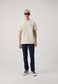 Cream-colored cotton t-shirt with "HUF" graphic, navy blue tapered pants, and gray sneakers with blue laces. Model wearing dark sunglasses.