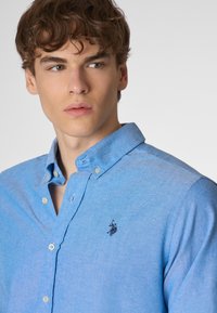 Camicia azzurro chiaro con bottoni, caratterizzata da una sottile trama, maniche lunghe, colletto classico e un logo ricamato a contrasto sul petto.