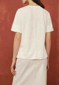Tricou alb texturat cu mâneci scurte și tiv rotunjit, având un detaliu cu o cusătură centrală pe spate, asortat cu o fustă deschisă.