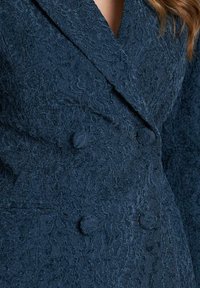 Blazer de encaje azul marino con un diseño de doble botonadura, tela texturizada y detalles de botones prominentes en la parte delantera.