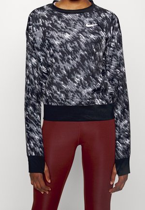Femme portant un haut court à manches longues Nike à motifs noirs et gris et un legging taille haute rouge foncé, debout devant un fond uni.