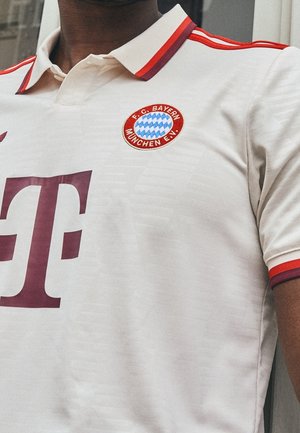 Vit F.C. Bayern München fotbollströja med röda och lila detaljer, stort kastanjefärgat "T"-logotyp och randig krage samt ärmsläpp.