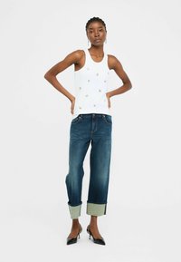 Top sin mangas blanco con bordados dorados pequeños, combinado con jeans de mezclilla oscuros con puños enrollados y ribete azul claro. Tacones altos negros.