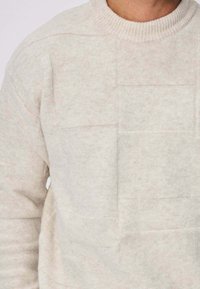 Pull en beige clair, texturé, avec un col rond côtelé. Présente un motif géométrique subtil et une apparence de tissu doux.