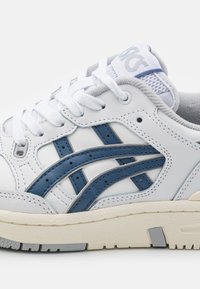 Baskets en cuir blanc avec des accents bleus, présentant trois rayures parallèles, une semelle texturée et des lacets blancs. La languette en mesh ajoute de la respirabilité.