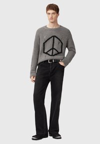 Giovane uomo che indossa un maglione grigio scolorito con un simbolo della pace nero, cintura nera, jeans neri e stivali neri, in piedi con una mano in tasca.