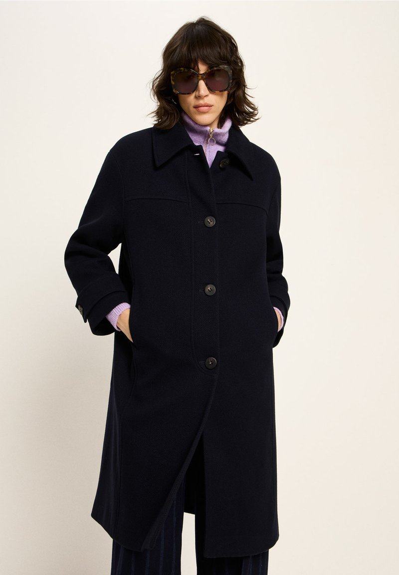 Navy wool coat with a collar, large buttons, and side pockets; paired with a light purple turtleneck and oversized tortoiseshell sunglasses.