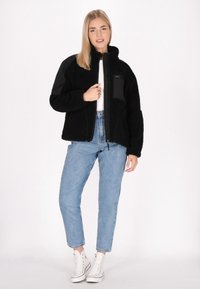 Giacca in pile nera con chiusura a zip frontale e collo alto, caratterizzata da un tessuto testurizzato, dettagli neri e un design aderente. Indossata con jeans in lavaggio chiaro.
