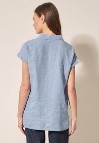 Lichtblauwe linnen korte mouwen overhemd met een kraag, met een relaxte pasvorm en opgerolde mouwen, weergegeven vanuit de achterkant tegen een beige achtergrond.