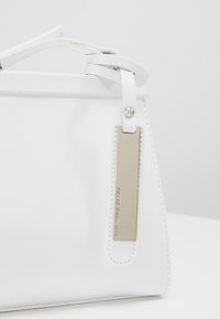 Biała syntetyczna torba na ramię o strukturze, z górnym uchwytem oraz metalową metką z wytłoczonym napisem "PIKADO SINCE 1913."