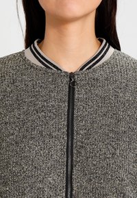 Grå texturerad bomberjacka med en svart dragkedja och randig ribbad krage. Tyget har ett blandat mönster och en figurnära design.