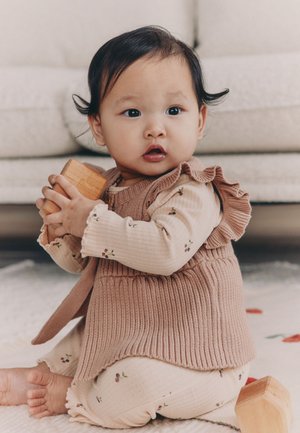 Taapero istuu lattialla ja pitelee puisia palikoita, pukeutuneena beigeen asuun, jossa on kukkakuviot ja neulottu liivi.