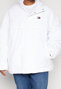 Hvid pufferjakke lavet af nylon, med lynlås-lukning, forlommer og et lille broderet logo på brystet. Glat tekstur.