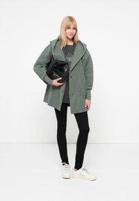 Grüne Fleecejacke mit Kapuze, Knopfverschluss, kombiniert mit einem dunkelgrauen Pullover und schwarzen Leggings, hält eine schwarze Tasche und trägt weiße Sneakers.