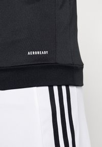Gros plan sur un haut de sport noir avec le texte "AEROREADY" au-dessus de l'ourlet côtelé, associé à un pantalon blanc avec trois bandes noires verticales.